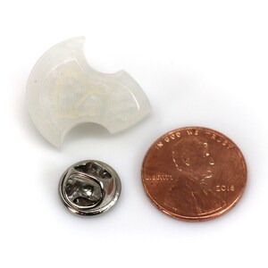 May include: A white, translucent, crescent-shaped pin with a silver pin back. The pin is about the size of a US penny.