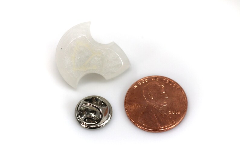 May include: A white, translucent, crescent-shaped pin with a silver pin back. The pin is about the size of a US penny.