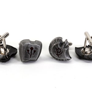 May include: Four silver cufflinks with different designs. Each cufflink features a shield-shaped design with a different symbol in the center. The symbols are a trident, a letter P, a shield, and a circle.