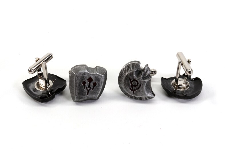 May include: Four silver cufflinks with different designs. Each cufflink features a shield-shaped design with a different symbol in the center. The symbols are a trident, a letter P, a shield, and a circle.