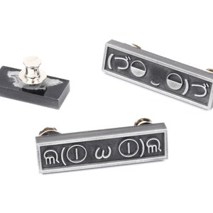 May include: Three silver-toned metal pins with different symbols. The pins are rectangular and have a black background. The symbols on the pins are: ( ) O ) , ( ) &omega; ( ) , and ( ) I &omega; ( ) .
