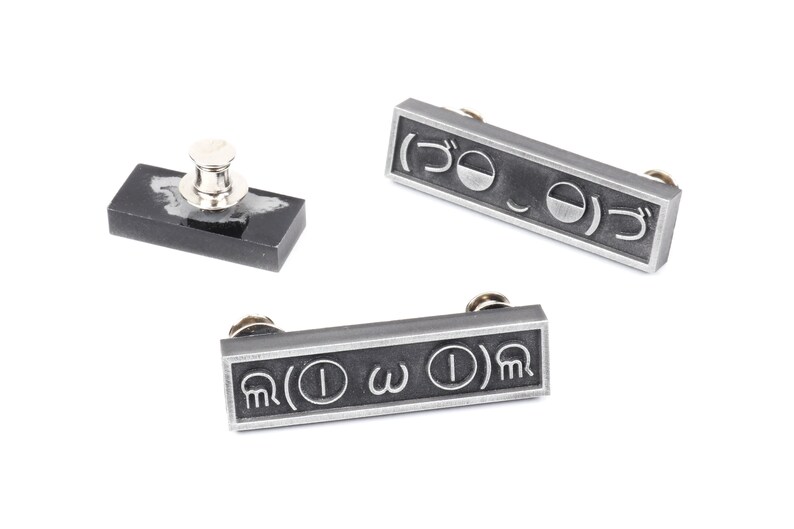 May include: Three silver-toned metal pins with different symbols. The pins are rectangular and have a black background. The symbols on the pins are: ( ) O ) , ( ) &omega; ( ) , and ( ) I &omega; ( ) .