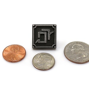K&ouml;nnte beinhalten: Ein quadratischer, silberfarbener Metallpin mit einem stilisierten Symbol in der Mitte, umgeben von einem Rahmen mit abgerundeten Ecken. Ein Cent, ein Dime und ein Viertel-Dollar sind zur Gr&ouml;&szlig;enveranschaulichung um den Pin herum platziert.