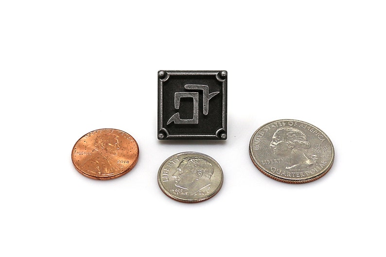 K&ouml;nnte beinhalten: Ein quadratischer, silberfarbener Metallpin mit einem stilisierten Symbol in der Mitte, umgeben von einem Rahmen mit abgerundeten Ecken. Ein Cent, ein Dime und ein Viertel-Dollar sind zur Gr&ouml;&szlig;enveranschaulichung um den Pin herum platziert.
