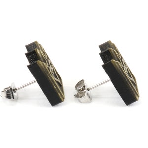May include: A pair of stud earrings with a rectangular design. The earrings have a dark, possibly black, base color with a gold-colored embossed pattern. The posts and backs are silver-colored.