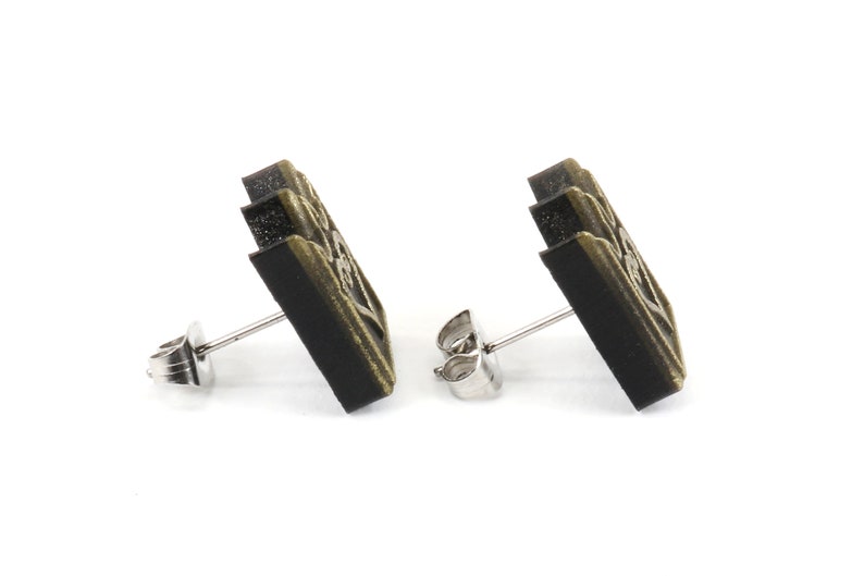 May include: A pair of stud earrings with a rectangular design. The earrings have a dark, possibly black, base color with a gold-colored embossed pattern. The posts and backs are silver-colored.