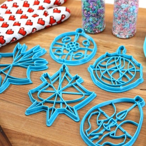 May include: A set of five blue plastic Christmas ornament cookie cutters on a wooden surface. The cutters are shaped like ornaments with different designs. There is a rolling pin, a spatula, and two jars of sprinkles in the background.