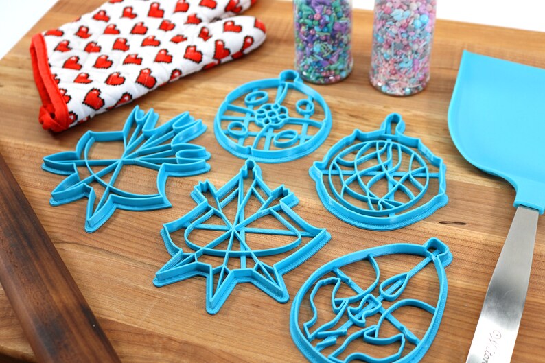 May include: A set of five blue plastic Christmas ornament cookie cutters on a wooden surface. The cutters are shaped like ornaments with different designs. There is a rolling pin, a spatula, and two jars of sprinkles in the background.