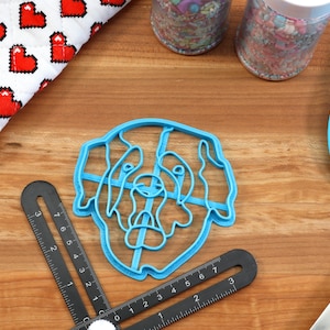 May include: A blue plastic dog-shaped cookie cutter, a blue spatula, and a black measuring tool are on a wooden surface. A red and white heart-patterned oven mitt and jars of sprinkles are also present. Baking and kitchen tools.