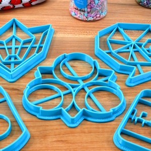 May include: Five blue plastic cookie cutters in different shapes, including a bottle, a flower, a geometric shape, and two different geometric star shapes. The cutters are on a wooden surface.