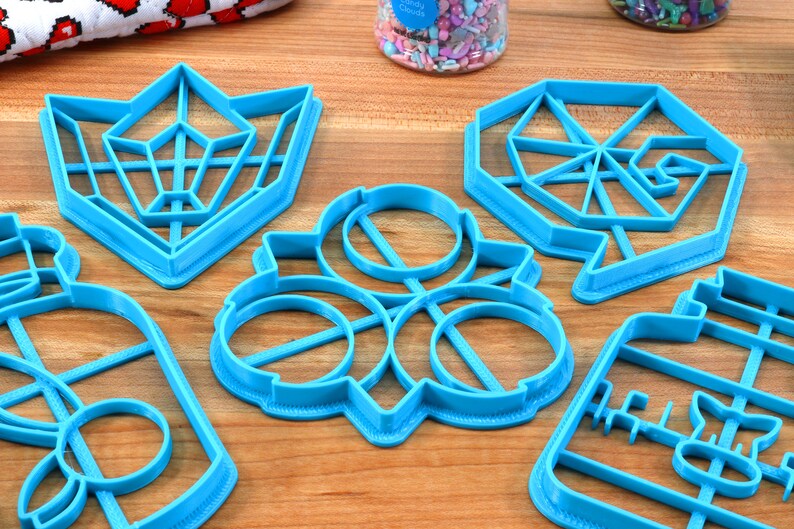 May include: Five blue plastic cookie cutters in different shapes, including a bottle, a flower, a geometric shape, and two different geometric star shapes. The cutters are on a wooden surface.