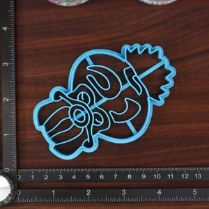May include: A blue plastic cookie cutter shaped like a cartoon character with a crown on its head. The character has a large nose and a wide mouth.