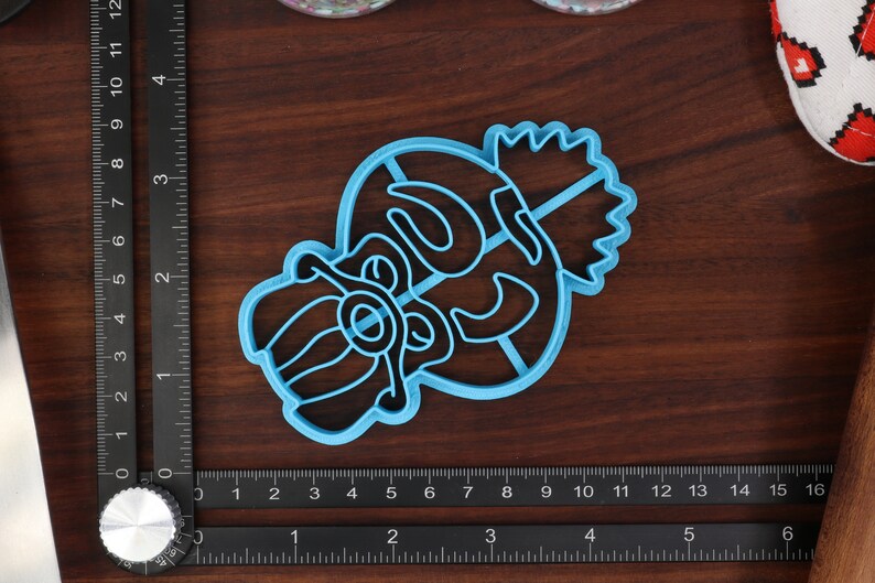 May include: A blue plastic cookie cutter shaped like a cartoon character with a crown on its head. The character has a large nose and a wide mouth.