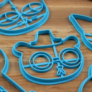 May include: A set of blue plastic cookie cutters shaped like cartoon characters. The cutters feature a bear wearing a hat and glasses, a cat with a bow tie, and a crown.