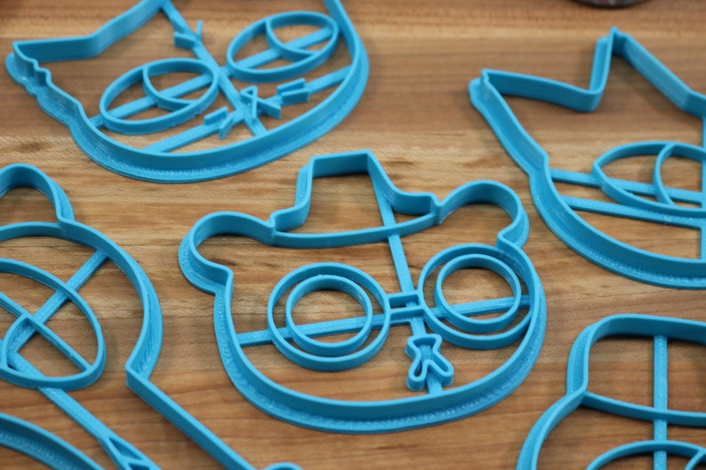 May include: A set of blue plastic cookie cutters shaped like cartoon characters. The cutters feature a bear wearing a hat and glasses, a cat with a bow tie, and a crown.