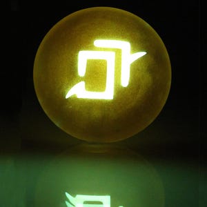 Może przedstawiać: Świecąca ż&oacute;łta kula z białym symbolem na niej. Symbol to stylizowana litera "D" z przechodzącą przez nią ukośną linią.