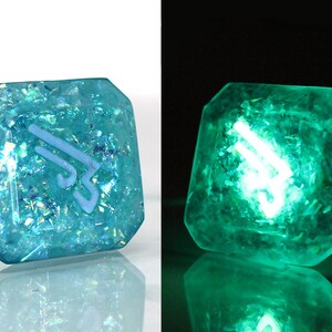 May include: A teal blue, square-shaped, resin dice with a white symbol and iridescent glitter embedded in the resin. The dice glows green in the dark.