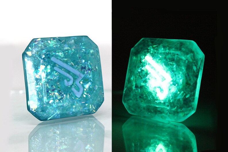 May include: A teal blue, square-shaped, resin dice with a white symbol and iridescent glitter embedded in the resin. The dice glows green in the dark.