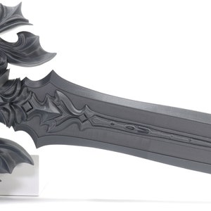 May include: A detailed, gray, metal sword with intricate carvings and a long, straight blade. The sword is on a white stand.