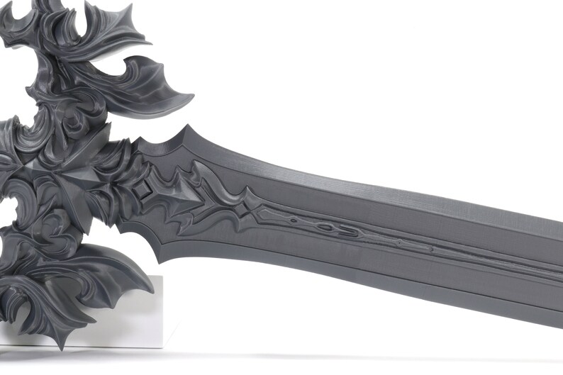 May include: A detailed, gray, metal sword with intricate carvings and a long, straight blade. The sword is on a white stand.