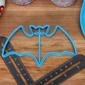 May include: A blue plastic bat-shaped cookie cutter on a wooden surface with a black ruler.