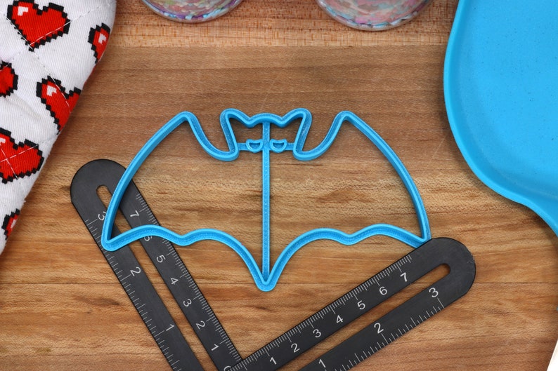 May include: A blue plastic bat-shaped cookie cutter on a wooden surface with a black ruler.