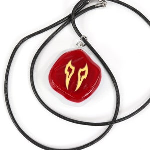 May include: A red and white pendant with a gold design hangs from a black cord necklace. The pendant is shaped like a rounded square and features two stylised flames in gold.