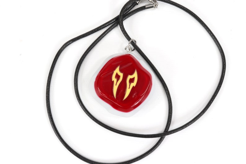 May include: A red and white pendant with a gold design hangs from a black cord necklace. The pendant is shaped like a rounded square and features two stylised flames in gold.