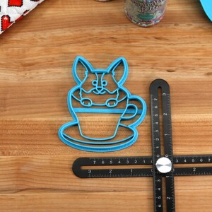 Pu&ograve; includere: Tagliapasta in plastica blu a forma di corgi seduto in una tazza da t&egrave;. Il tagliapasta &egrave; su una superficie di legno con un righello nero.