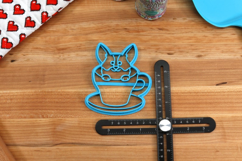 Pu&ograve; includere: Tagliapasta in plastica blu a forma di corgi seduto in una tazza da t&egrave;. Il tagliapasta &egrave; su una superficie di legno con un righello nero.