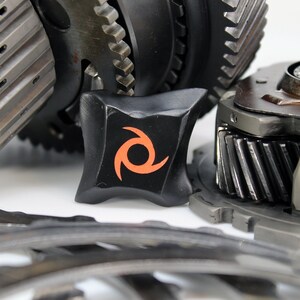 May include: Close-up of a black gear with an orange swirl logo. The gear is surrounded by other gears and metal parts.