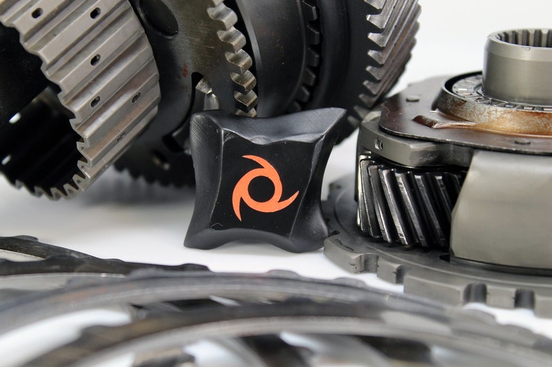 May include: Close-up of a black gear with an orange swirl logo. The gear is surrounded by other gears and metal parts.