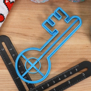 May include: A blue plastic key-shaped cookie cutter with a circular cutout in the centre. The cutter is on a wooden surface with a black ruler.