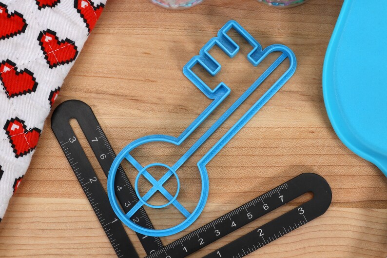 May include: A blue plastic key-shaped cookie cutter with a circular cutout in the centre. The cutter is on a wooden surface with a black ruler.