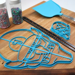 May include: A set of blue plastic cookie cutters shaped like a video game character, a skull, and a spatula. The cutters are on a wooden surface with two jars of sprinkles.