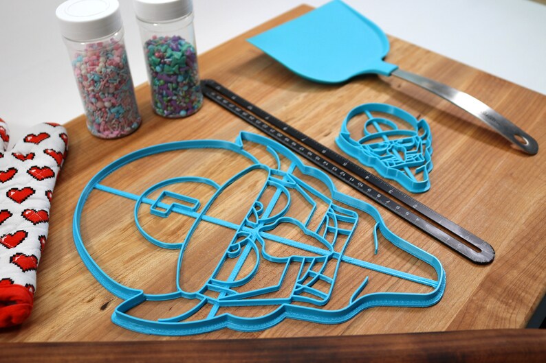 May include: A set of blue plastic cookie cutters shaped like a video game character, a skull, and a spatula. The cutters are on a wooden surface with two jars of sprinkles.
