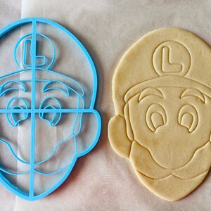 May include: A blue plastic cookie cutter shaped like a cartoon character's face, next to a cut-out cookie. The cookie cutter has a detailed design, including the character's hat with an "L" on it, eyes, and mustache. The cookie is light brown.