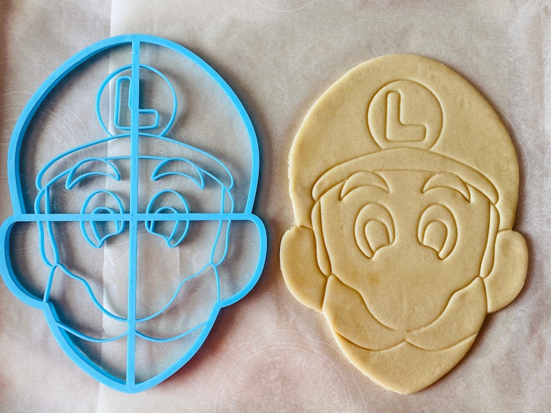 May include: A blue plastic cookie cutter shaped like a cartoon character's face, next to a cut-out cookie. The cookie cutter has a detailed design, including the character's hat with an "L" on it, eyes, and mustache. The cookie is light brown.