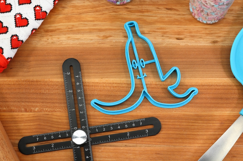 May include: A blue plastic banana-shaped cookie cutter with a smiling face and two eyes. The cutter is on a wooden surface.