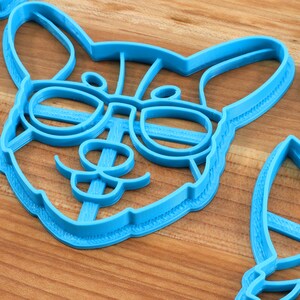 Pu&ograve; includere: Un tagliapasta di plastica blu a forma di cane con gli occhiali. Il cane ha i baffi e un'espressione felice.
