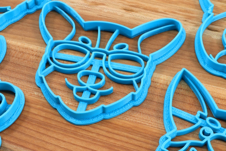 Pu&ograve; includere: Un tagliapasta di plastica blu a forma di cane con gli occhiali. Il cane ha i baffi e un'espressione felice.