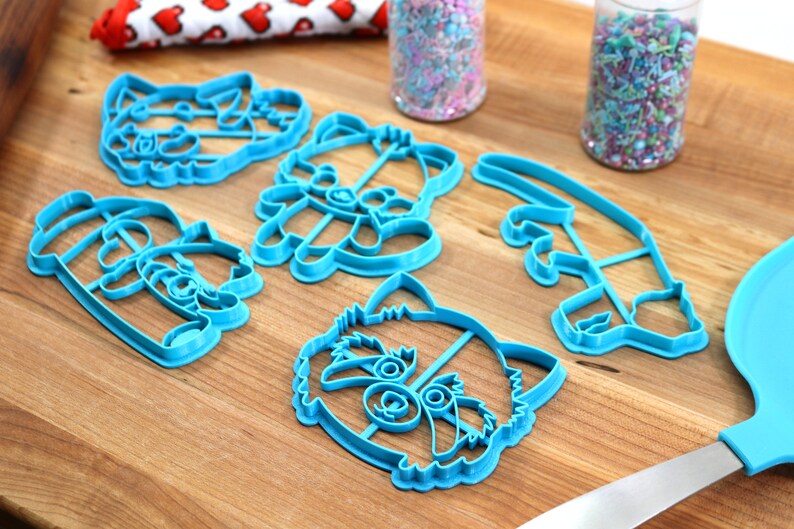 May include: Set of six blue plastic cookie cutters shaped like a raccoon, including a full face, a head, a tail, and two different poses of the raccoon holding a stick.
