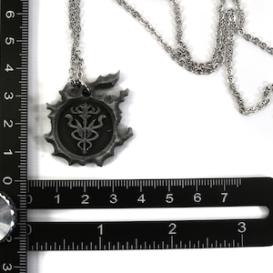 May include: A silver necklace with a pendant featuring a detailed, circular design with a stylized symbol inside. The pendant is about 2 inches in diameter and is measured against a ruler.