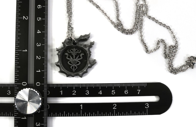 May include: A silver necklace with a pendant featuring a detailed, circular design with a stylized symbol inside. The pendant is about 2 inches in diameter and is measured against a ruler.