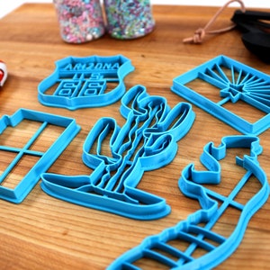 May include: A set of six blue plastic cookie cutters shaped like a cactus, a state of Arizona road sign, a window, a cowboy boot, a horse, and the Arizona state flag.