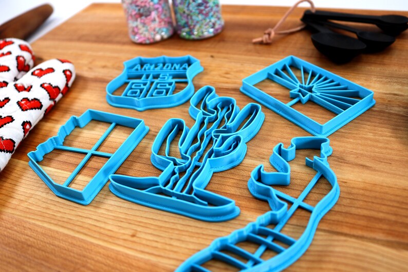 May include: A set of six blue plastic cookie cutters shaped like a cactus, a state of Arizona road sign, a window, a cowboy boot, a horse, and the Arizona state flag.
