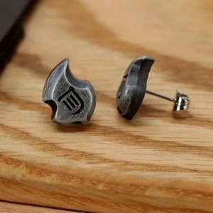 May include: A pair of silver-colored stud earrings. Each earring has a unique, stylized shape with a curved top and a small, engraved design. The earrings are set against a wooden background.