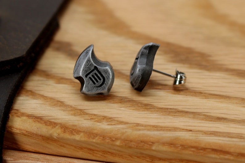 May include: A pair of silver-colored stud earrings. Each earring has a unique, stylized shape with a curved top and a small, engraved design. The earrings are set against a wooden background.