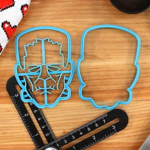 May include: Two blue plastic cookie cutters, one shaped like a Frankenstein's monster face and the other shaped like a generic head. The cutters are on a wooden surface.