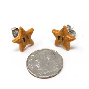 May include: A pair of gold star-shaped earrings with black eyes. The earrings are made of polymer clay and have a shiny finish. The earrings are about the size of a dime.